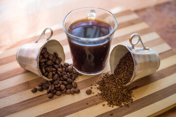 On a wooden plank in the middle is a cup of brewed coffee, the other two cups are filled with whole, roasted beans and instant coffee, and they are scattered on the plank.