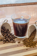 On a wooden plank in the middle is a cup of brewed coffee, the other two cups are filled with whole, roasted beans and instant coffee, and they are scattered on the plank.