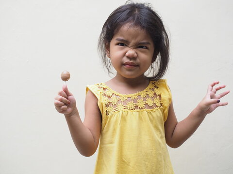 A Cute Asian Kid Girl Wearing Yellow Shirt Showing Silly Face Expression While Holding A Lollipop Isolated On White. Space For Copy