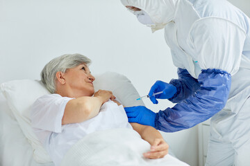 Modern doctor wearing protective suit giving injection to mature woman lying on bed in hospital ward