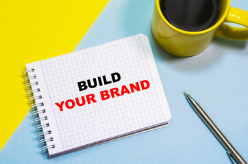 BUILD YOUR BRAND on a notepad on a blue-yellow background, a cup of coffee and a pen. Business concept