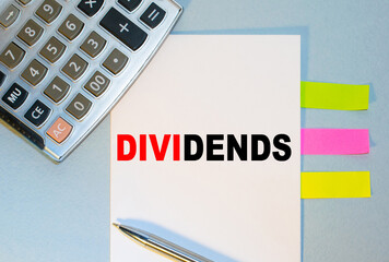 DIVIDENDS is written on a notepad next to multicolored stickers, a calculator and a pen on a blue background. Business concept