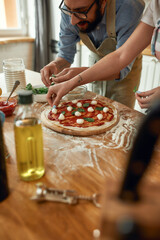 Young couple making pizza together at home, man in apron, professional cook adding basil on the...
