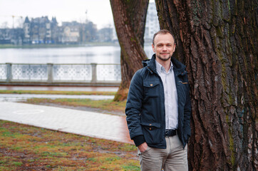 Handsome businessman freelancer in blue dark coat and beige trousers smiles and standing near tree