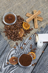 Coffee beans on rough linen and candle stub. Ground coffee in containers and spices. On a surface of brushed pine boards painted black and white.