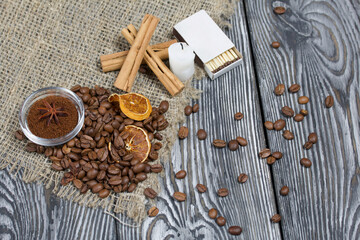 Coffee beans on rough linen and candle stub. Ground coffee in a container and spices. On a surface of brushed pine boards painted black and white.