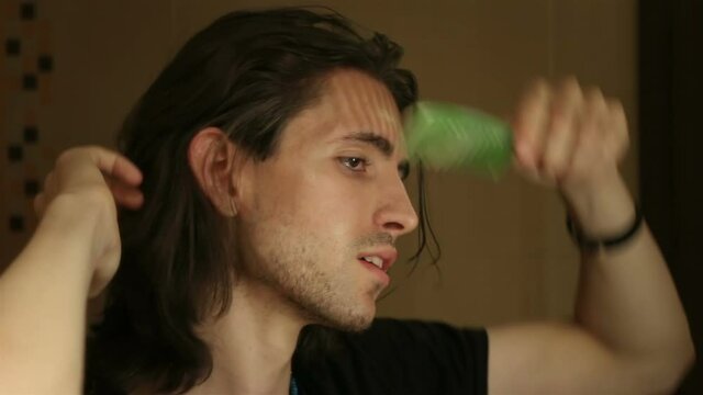 Portrait of young man brushing long layered dark brown hair in bathroom, standing in front of mirror, face profile, detail