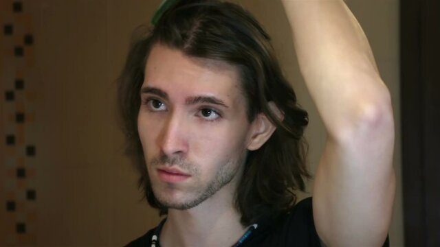 Young man brushing long layered dark brown hair in bathroom standing in front of mirror, face detail