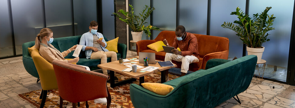 Business team during the COVID-19 outbreak, young multiracial office workers wearing medical protective masks working together while sitting in lounge zone in coworking office