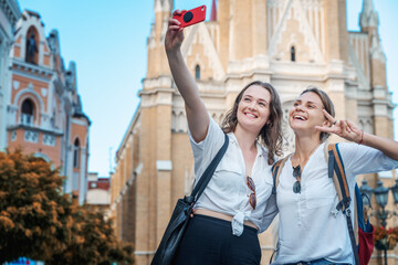 Fototapeta premium Two young happy pretty female students taking selfie on a mobile phone against the background of a european city