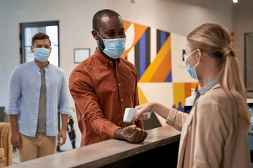 Young woman wearing medical face mask standing at reception in the office and using infrared...