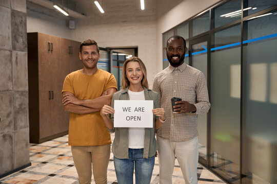 Young Happy Woman, Female Office Worker Showing Paper With Text WE ARE OPEN At Camera And Smiling While Standing With Her Colleagues In The Modern Office