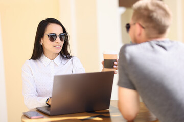 Woman in sunglasses at table with laptop and coffee maker communicates with man. Business communication in cafe concept