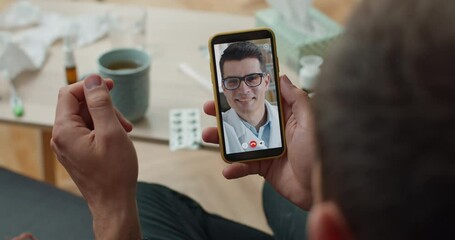 Overshoulder view of young male doctor consulting patient by video call on smartphone and writing details. Over shoulder closeup mobilephone screen view. Concept of telemedicine