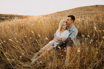 Naklejka na ściany i meble Young couple of man and woman sitting in a golden field, hugging.