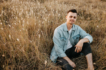 Naklejka na ściany i meble Young handsome man, wearing denim jacket, sitting in a field, looking to camera.