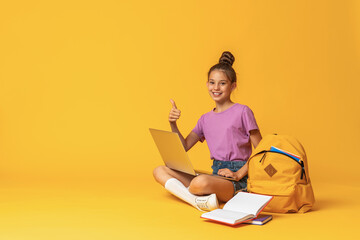 happy schoolgirl with a backpack, laptop and books poses on a pink background.