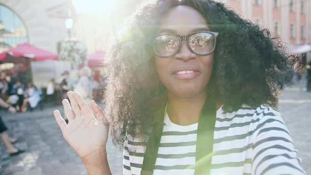 Close up of beautiful African American woman standing in city center and waving hand at camera. Happy girl recording video outside in sunlight street. Camera view.