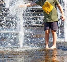 A teenager stands barefoot by a fountain and tries to stop a powerful stream of water with his hand on a bright hot day.