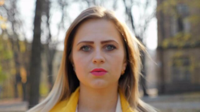 Close up woman in yellow cloak, white blouse stands and looks at the camera in the park, camera moves girl. Girl with pink lipstick on lips outdoors. 4K slow motion footage