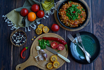 Cutting board with tomatoes, garlic, corn, hot pepper and a knife, serving plates and two forks, stewed vegetables in a pan, olive oil, beans and vegetables on a dark wooden background