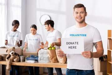 Selective focus of young volunteer holding card with donate here lettering in charity center