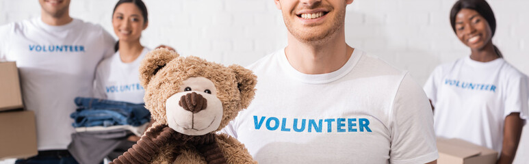 Panoramic shot of man holding soft toy near multiethnic volunteers in charity center