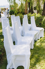 Chairs with white cloth stand in a row on the green grass at a wedding ceremony.