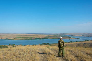 Man traveler in hat on mountain top looks at river below against blue sky. Copy space, soft focus. Freedom and travel concept