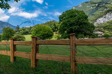 Fototapeta premium Tenno lake, a beautiful summer panorama of the green meadows and the mountains of Trentino. Delicate clouds. Wooden fence near the shore. This lake is close to Garda lake, Trentino Alto Adige, Italy.