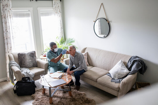 Home Caregiver In Face Mask Taking Temperature Of Senior Man On Sofa