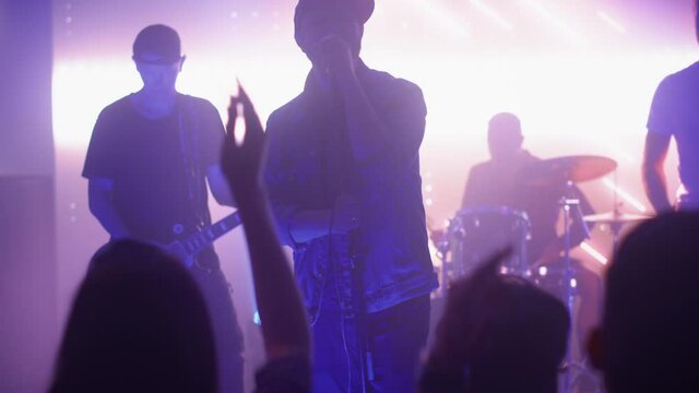 Rock Band with Guitarists and Drummer Performing at a Concert in a Night Club. Front Row Crowd is Partying. Silhouettes of Fans Raise Hands in Front of Bright Colorful Strobing Lights on Stage. 