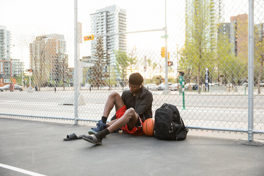 Young Man Putting On Basketball Shoes At Fence Of Urban Court