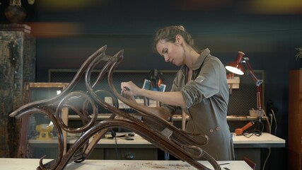 Restoration of wooden rocking chair in a workshop. A working female carpenter peels off paint from...