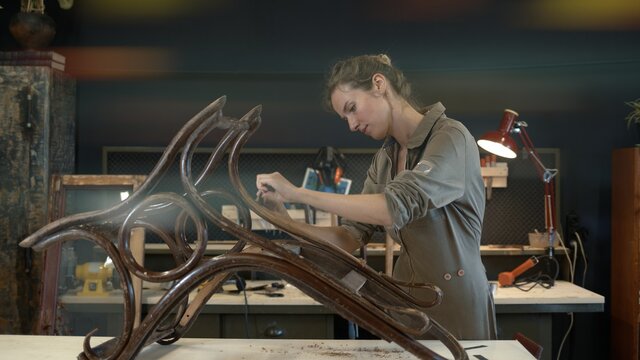 Restoration of wooden rocking chair in a workshop. A working female carpenter peels off paint from rocking chair legs with a spatula, restorer in a workshop, Lense flare