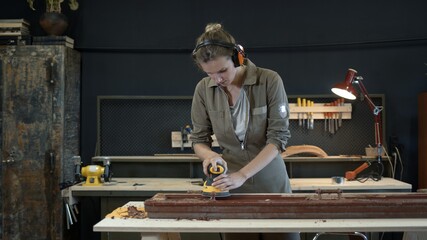 Young woman grinds the wooden door, restoration works. Female wearing hearing protection device grinding, polishing old door in workshop