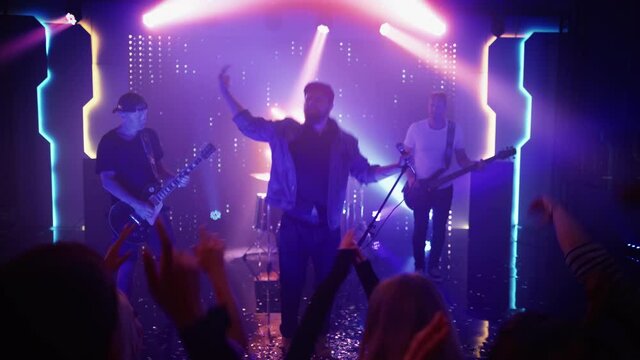 Rock Band with Guitarists and Drummer Performing at a Concert in a Night Club. Front Row Crowd is Partying. Silhouettes of Fans Raise Hands in Front of Bright Colorful Strobing Lights on Stage. 