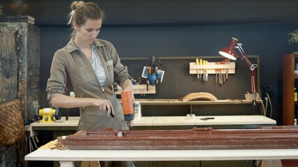 Restoration of wooden doors, window. A working female in worksuit carpenter peels off paint from a wooden product with a spatula and a heat gun, lense flare