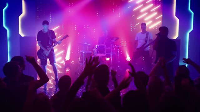 Rock Band with Guitarists and Drummer Performing at a Concert in a Night Club. Front Row Crowd is Partying. Silhouettes of Fans Raise Hands in Front of Bright Colorful Strobing Lights on Stage. 
