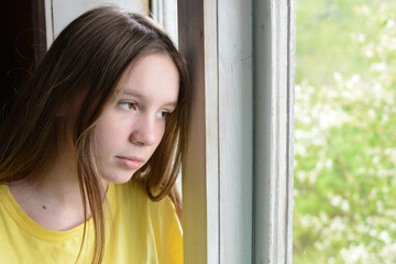 A cute brown-eyed girl in a yellow T-shirt with a sad expression looks out the window. Close-up, horizontal photo.