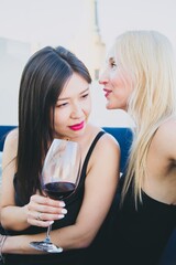 A blonde girl privately talks to an Asian girl in a cocktail dress with a glass of red wine in a party. Small talk between two girls at a social event.