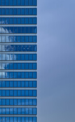 A tall mirrored skyscraper reflecting a blue sky with a cloud in it beside a view of a gray overcast sky