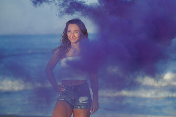 Beautiful young brunette woman in white shirt and denim shorts poses on beach - with purple smoke in air near her