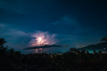 Lightnings, stars and dramatic clouds in the night sky over valley with trees, sea,  city and mountains. View from the top.