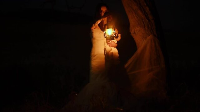 Scary woman with a lantern in night scene - Spooky image of a scary woman with dark eyes and appearance of a witch, in a white dress, holding a lit lantern and a frightening doll, in a dark night
