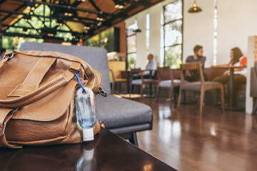 Mini portable alcohol gel bottle to kill Corona Virus(Covid-19) hang on a brown leather shoulder bag on table in coffee shop.New normal lifestyle. Health care concept. Selective focus on alcohol gel