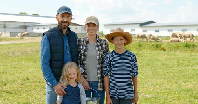 Portrait of Caucasian happy mother and father with little kids standing at pasture with sheep flock on background and smiling to camera. Joyful parents with small son and daughter in field at farm.