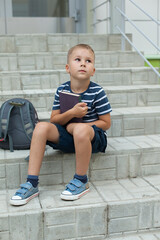 A small boy sits on the steps of the school, holding notebooks and looking up at the sky. Change.