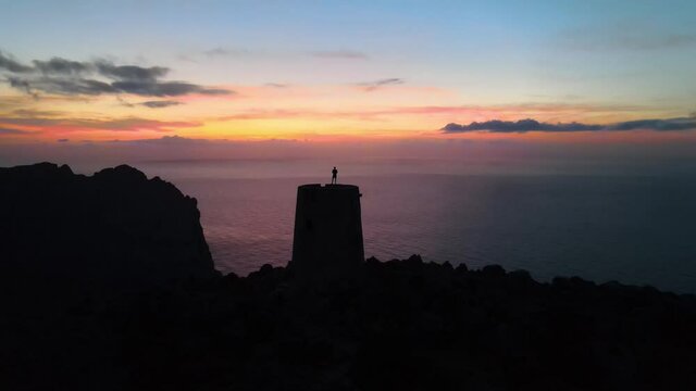 Aerial shot flying over the silhouette of a man standing on top of a watchtower on a spectacular sunset