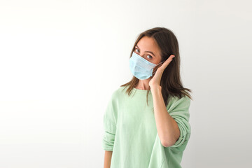 Adult female in medical mask keeping palm near ear and looking at camera while pretending to make call during pandemic against gray background